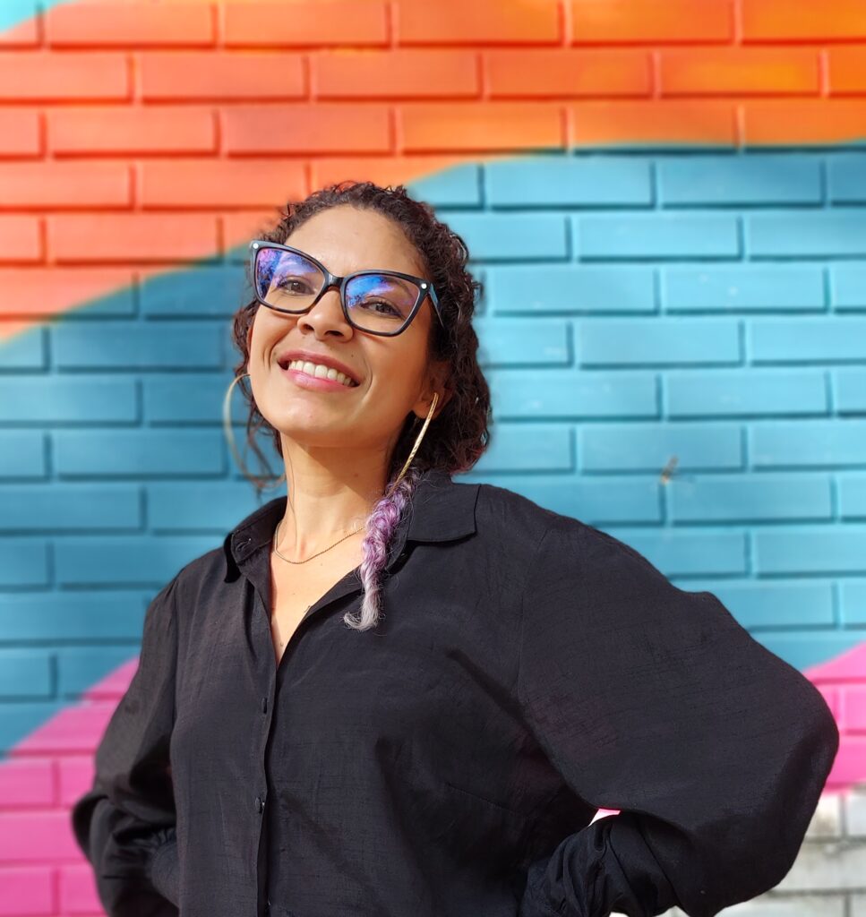foto da autora do texto com camisa preta e óculos sorrindo em fundo colorido
